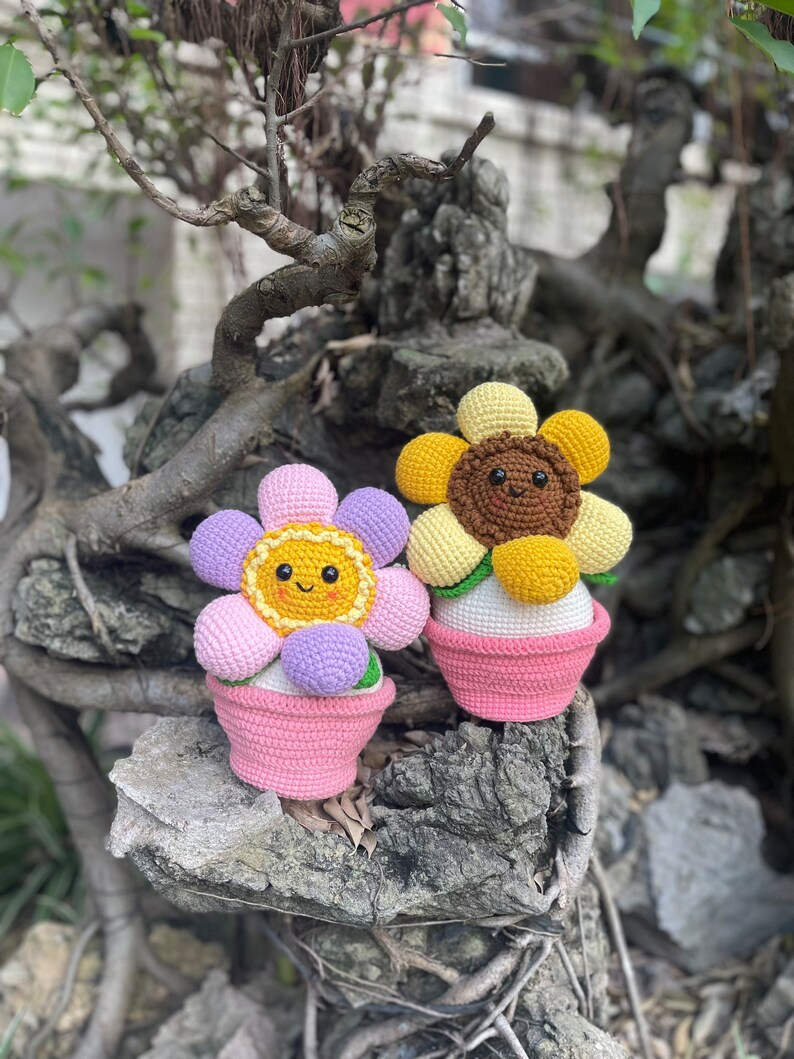 May include: Two crocheted flower pot decorations. One is a pink and purple flower with a yellow center and a pink pot. The other is a yellow flower with a brown center and a pink pot.