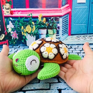 May include: A crocheted green and brown turtle with a white and yellow flower pattern on its shell. The turtle is being held in a person's hands.
