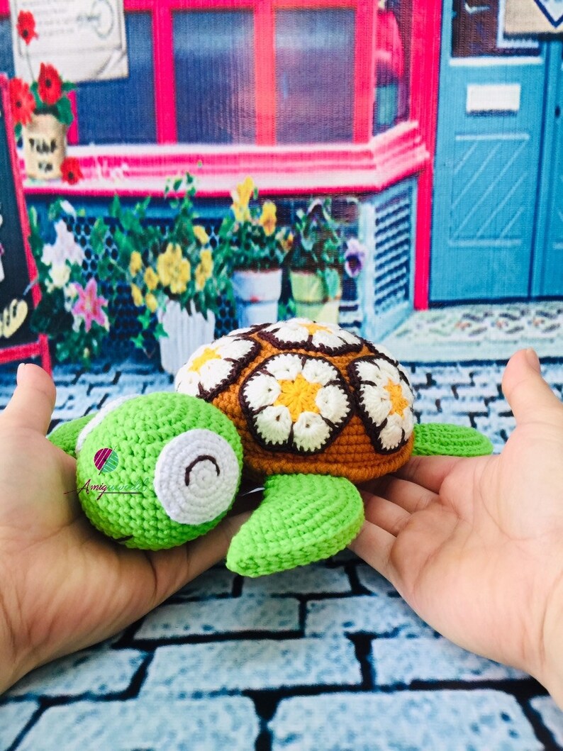 May include: A crocheted green and brown turtle with a white and yellow flower pattern on its shell. The turtle is being held in a person's hands.