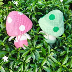 May include: Two crocheted mushroom plushies, one pink with white dots and one green with green dots, are sitting in a bed of green leaves.