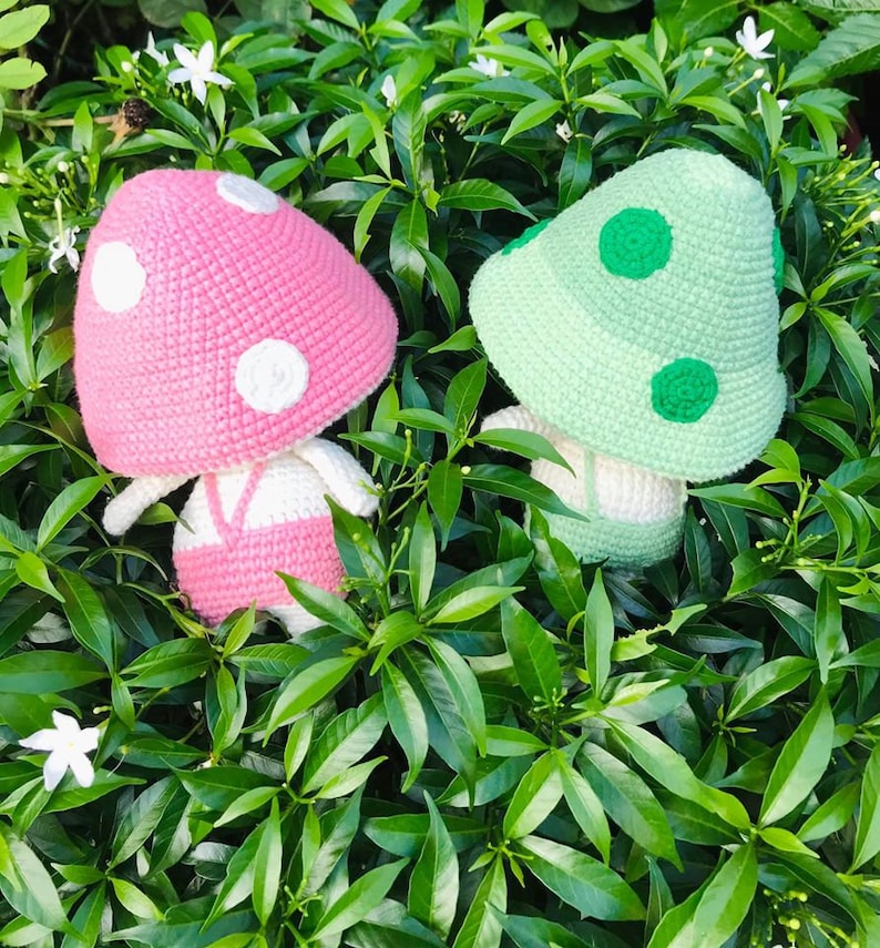 May include: Two crocheted mushroom plushies, one pink with white dots and one green with green dots, are sitting in a bed of green leaves.