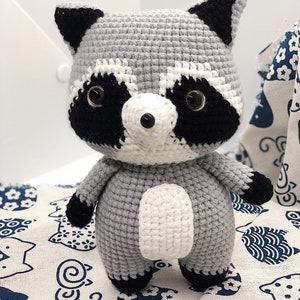 May include: A crocheted raccoon plush toy with black ears, eyes, and nose, and a white belly. The toy is standing on a white fabric with a blue and white pattern.