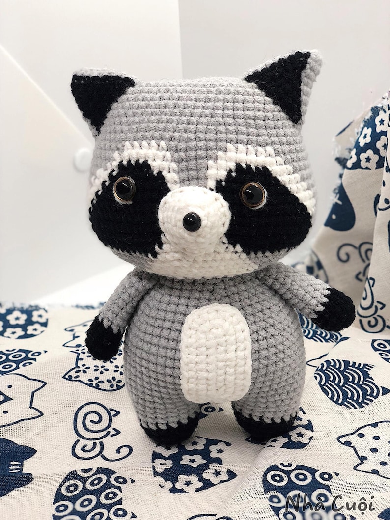 May include: A crocheted raccoon plush toy with black ears, eyes, and nose, and a white belly. The toy is standing on a white fabric with a blue and white pattern.