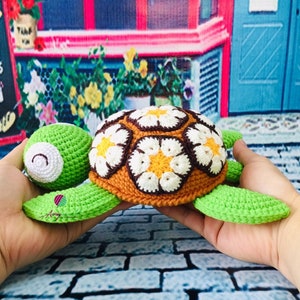 May include: A crocheted turtle toy with a brown, white, and orange shell and green limbs. The shell is decorated with a daisy pattern.