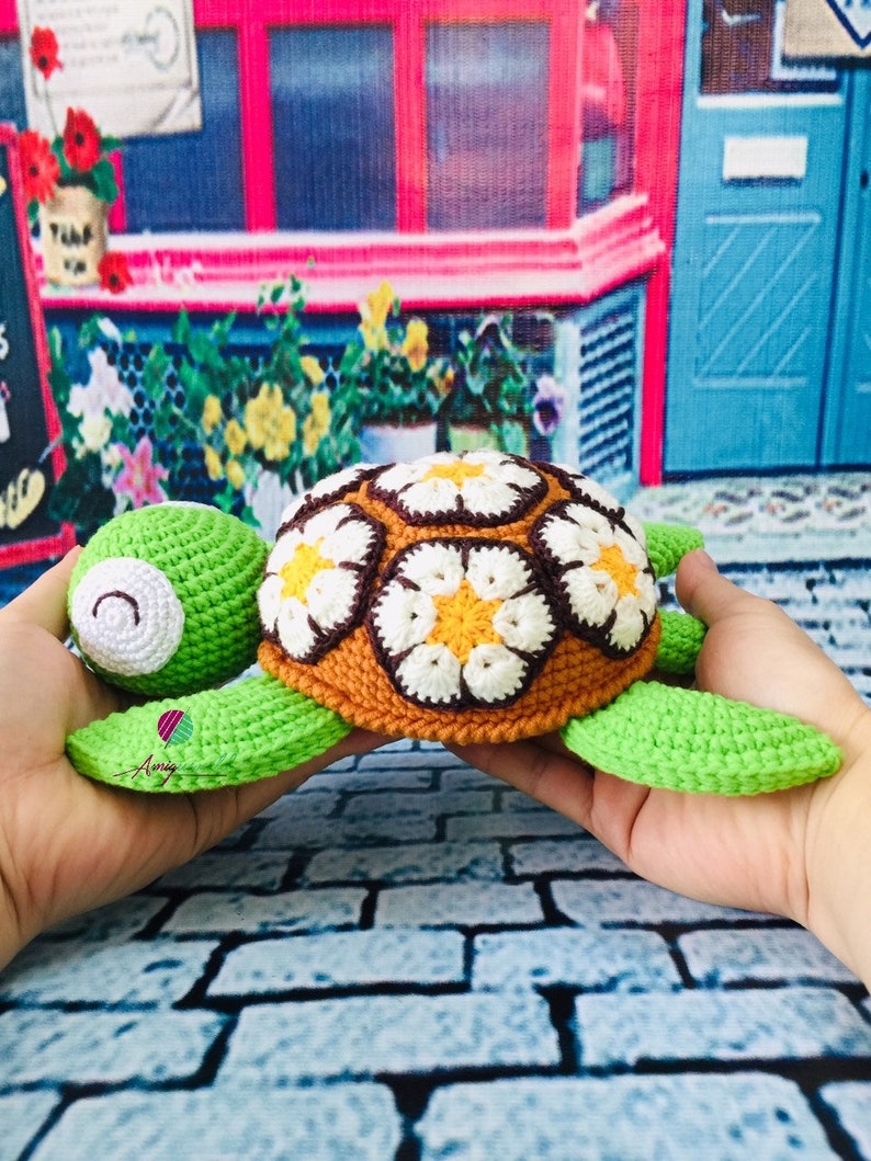 May include: A crocheted turtle toy with a brown, white, and orange shell and green limbs. The shell is decorated with a daisy pattern.