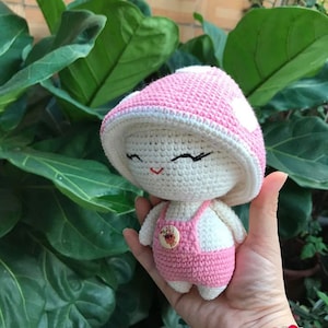 May include: A crocheted mushroom doll with a pink and white hat, white body, and pink overalls. The doll has a button on the front of its overalls.