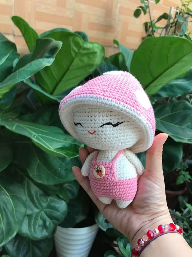 May include: A crocheted mushroom doll with a pink and white hat, white body, and pink overalls. The doll has a button on the front of its overalls.