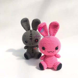 May include: Two crocheted bunny plushies, one grey with stitched details and one pink with embroidered eyes and a stitched mouth.
