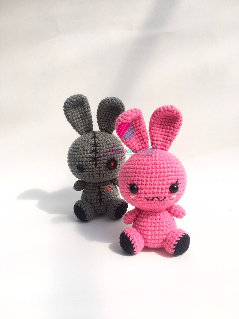 May include: Two crocheted bunny plushies, one grey with stitched details and one pink with embroidered eyes and a stitched mouth.