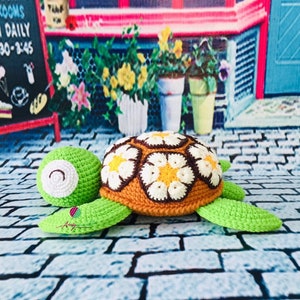 May include: A crocheted turtle toy with a brown shell decorated with white and yellow flowers. The turtle is green and has a white face with black eyes. The toy is sitting on a brick surface.