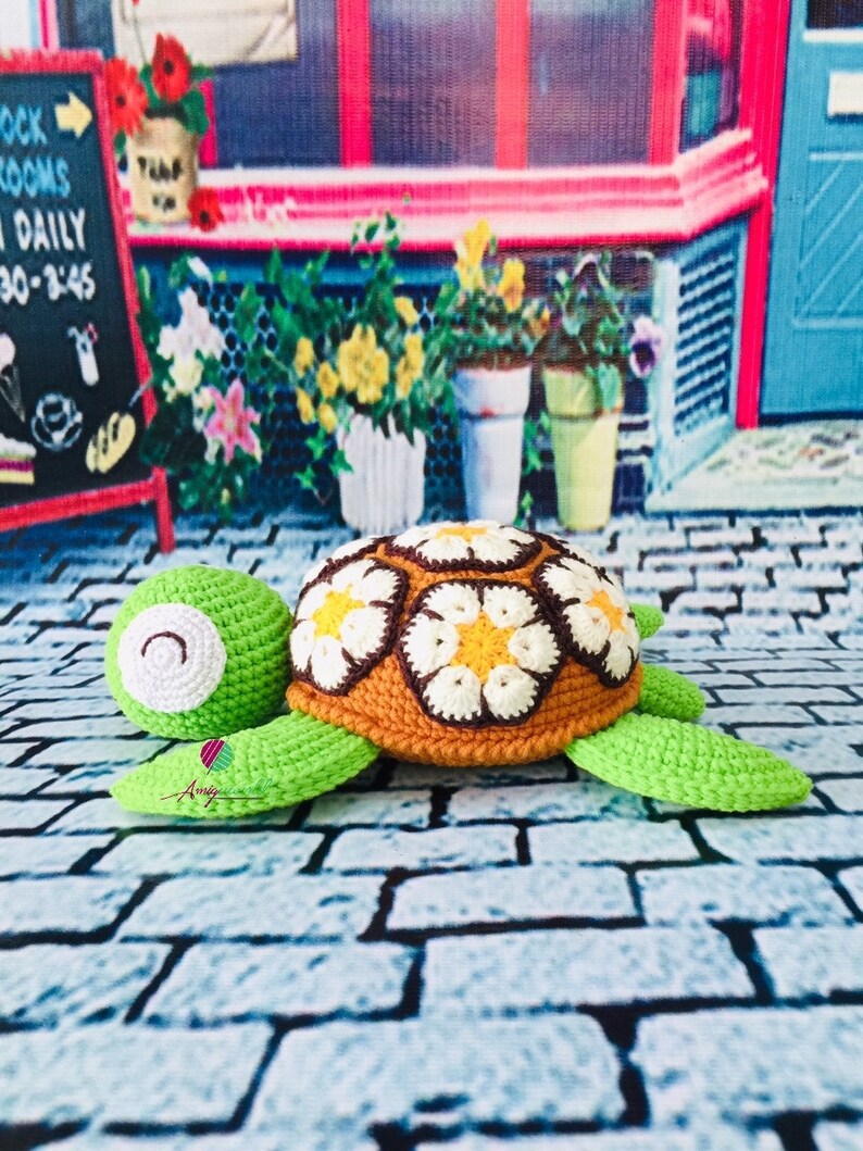 May include: A crocheted turtle toy with a brown shell decorated with white and yellow flowers. The turtle is green and has a white face with black eyes. The toy is sitting on a brick surface.