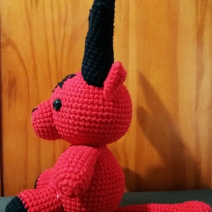 May include: A red crocheted plush toy with black horns and a tail. The toy is sitting on a dark surface.