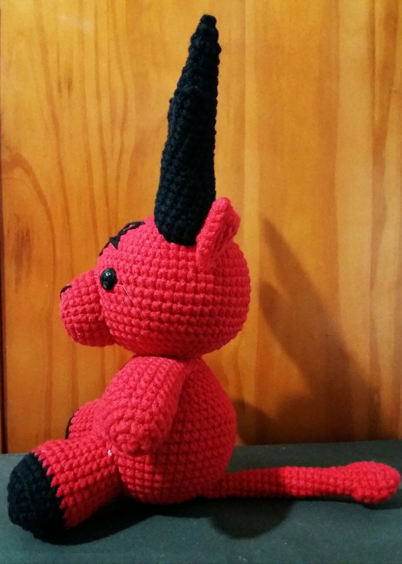 May include: A red crocheted plush toy with black horns and a tail. The toy is sitting on a dark surface.