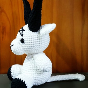 May include: A white crocheted stuffed animal with black horns, a black tail, and black feet. The animal has a black pentagram on its forehead.
