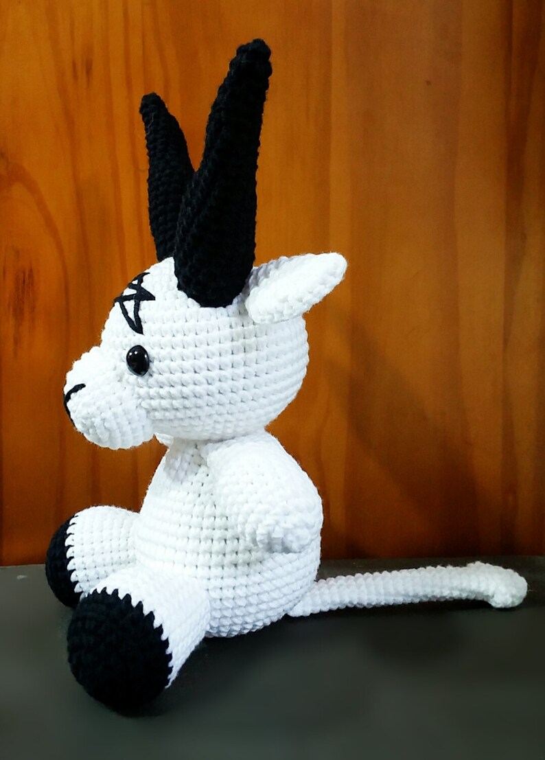 May include: A white crocheted stuffed animal with black horns, a black tail, and black feet. The animal has a black pentagram on its forehead.