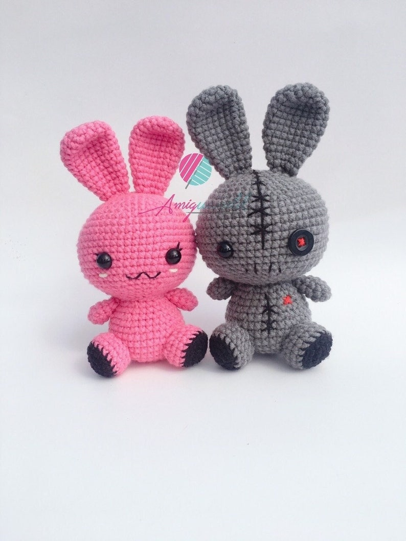 May include: Two crocheted bunny plushies, one pink and one grey. The pink bunny has a cute face with black eyes and a stitched smile. The grey bunny has a stitched-up face with a black button eye and a black button nose.