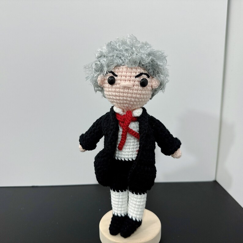 May include: A crocheted doll with gray hair, a black suit, a red bow tie, and white pants. The doll is standing on a wooden base.
