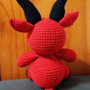 May include: A red crocheted devil plush toy with black horns and a tail. The toy is sitting on a gray surface.