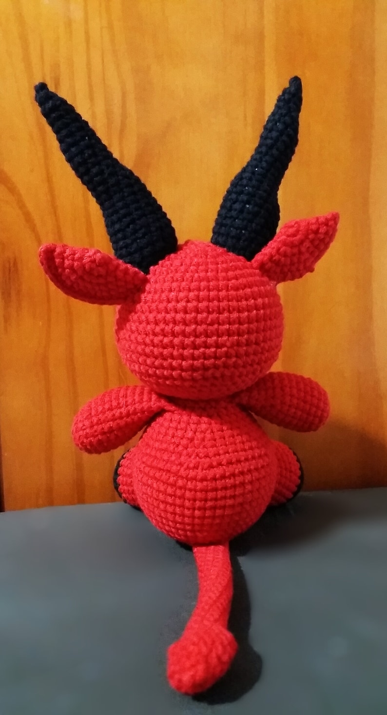 May include: A red crocheted devil plush toy with black horns and a tail. The toy is sitting on a gray surface.