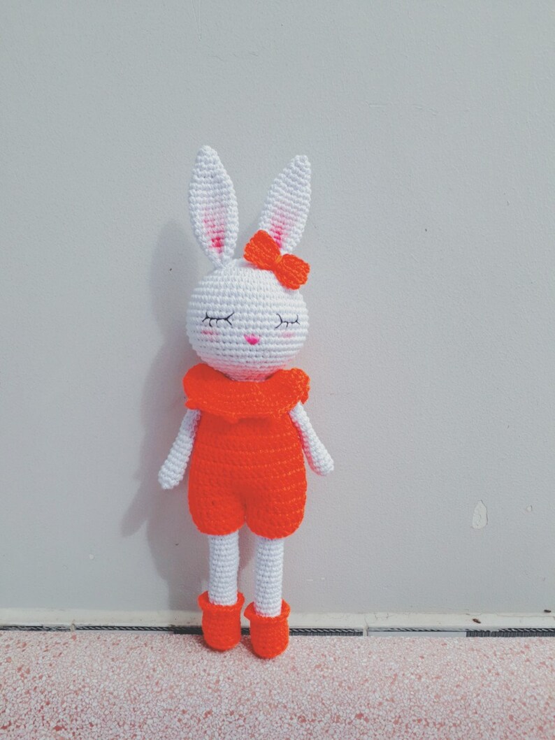 May include: White crocheted bunny with orange overalls and a red bow. The bunny has closed eyes and a pink nose.