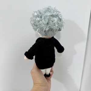 May include: A crocheted doll with gray hair, wearing a black jacket and white pants.