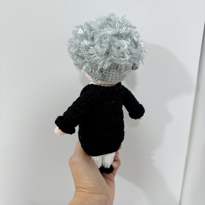 May include: A crocheted doll with gray hair, wearing a black jacket and white pants.