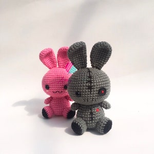May include: Two crocheted bunny plushies, one pink and one grey. The grey bunny has a stitched-up mouth and a single red button eye.
