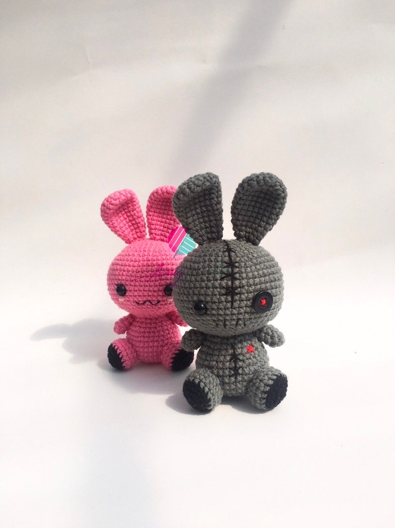 May include: Two crocheted bunny plushies, one pink and one grey. The grey bunny has a stitched-up mouth and a single red button eye.