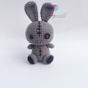 May include: A grey crocheted bunny with black button eyes and a red heart on its chest. The bunny has stitched seams and is sitting on a white background.