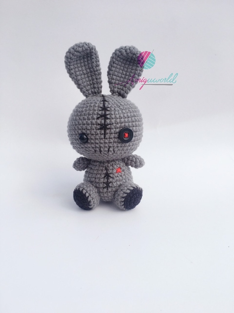 May include: A grey crocheted bunny with black button eyes and a red heart on its chest. The bunny has stitched seams and is sitting on a white background.