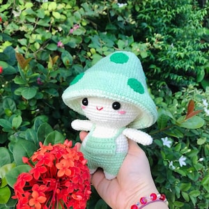 May include: A crocheted mushroom-shaped plush toy with a green hat and white body. The toy is wearing green overalls and has a happy face.