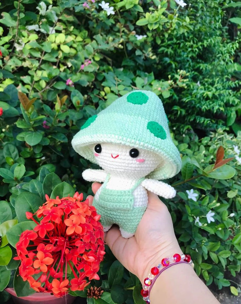 May include: A crocheted mushroom-shaped plush toy with a green hat and white body. The toy is wearing green overalls and has a happy face.