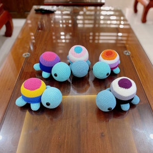 May include: Six crocheted turtle toys with different coloured shells. The turtles are all blue with black eyes and are arranged on a wooden surface.