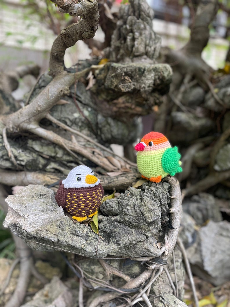 May include: Two crocheted bird toys, one brown and white with a yellow beak and the other green, orange, and red with a yellow beak, are perched on a tree branch.