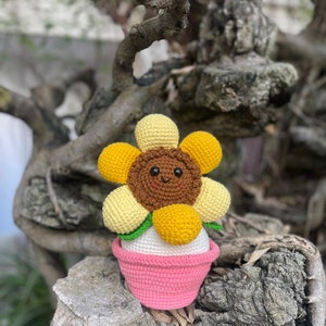 May include: A crocheted sunflower with a brown face and yellow petals is sitting in a pink pot. The sunflower has a happy face and is sitting on a tree branch.