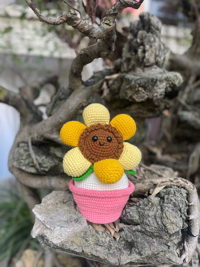 May include: A crocheted sunflower with a brown face and yellow petals is sitting in a pink pot. The sunflower has a happy face and is sitting on a tree branch.