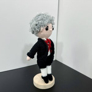 May include: A crocheted doll wearing a black suit with a red bow tie and white pants. The doll has gray hair and is standing on a wooden base.