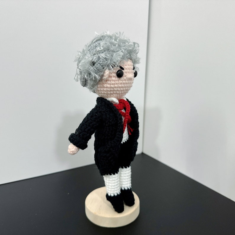 May include: A crocheted doll wearing a black suit with a red bow tie and white pants. The doll has gray hair and is standing on a wooden base.