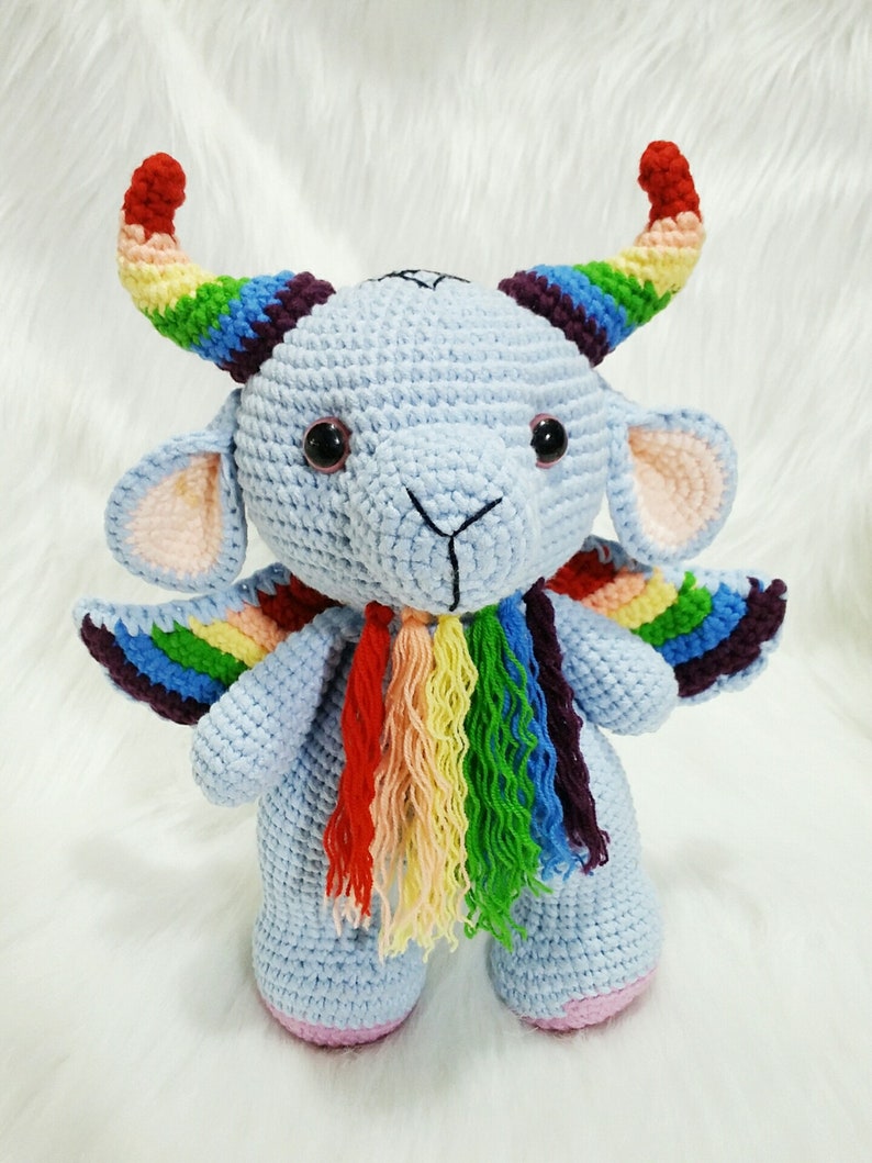 May include: A blue crocheted stuffed animal with rainbow horns, wings, and a scarf. The animal has large eyes and a small, pink nose.