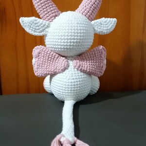May include: A white crocheted stuffed animal with pink ears, wings, and a tail. The animal has a rounded body and a long tail with a pink tassel.