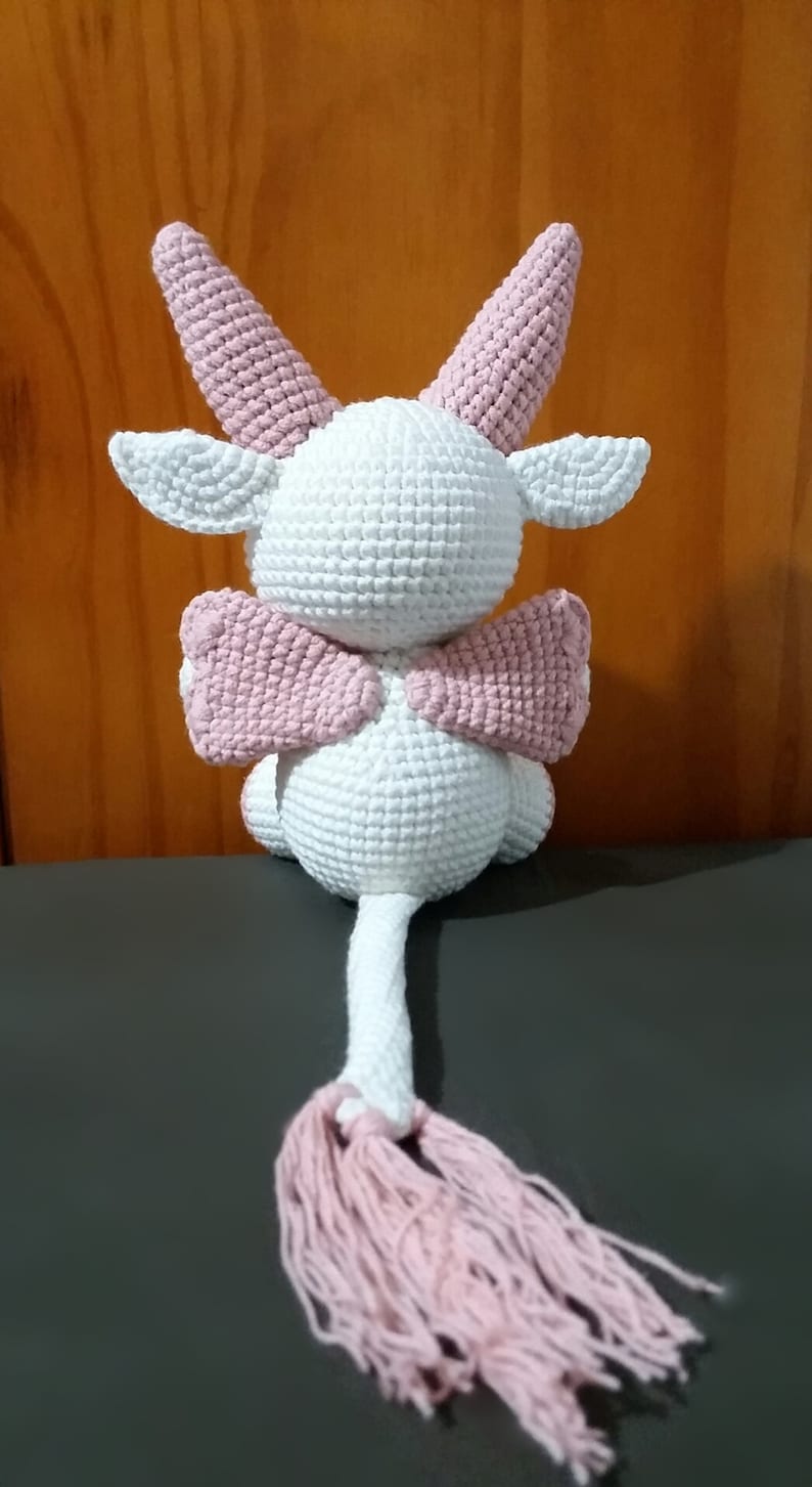 May include: A white crocheted stuffed animal with pink ears, wings, and a tail. The animal has a rounded body and a long tail with a pink tassel.
