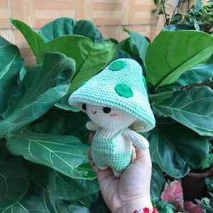 May include: A crocheted green mushroom-shaped plush toy with white accents and a cute face. The toy is being held in a hand with a red beaded bracelet.