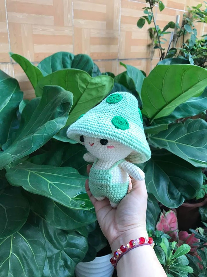 May include: A crocheted green mushroom-shaped plush toy with white accents and a cute face. The toy is being held in a hand with a red beaded bracelet.