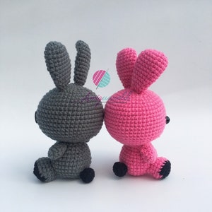 May include: Two crocheted bunny plushes, one grey and one pink, sitting back to back. They have black eyes and black feet.