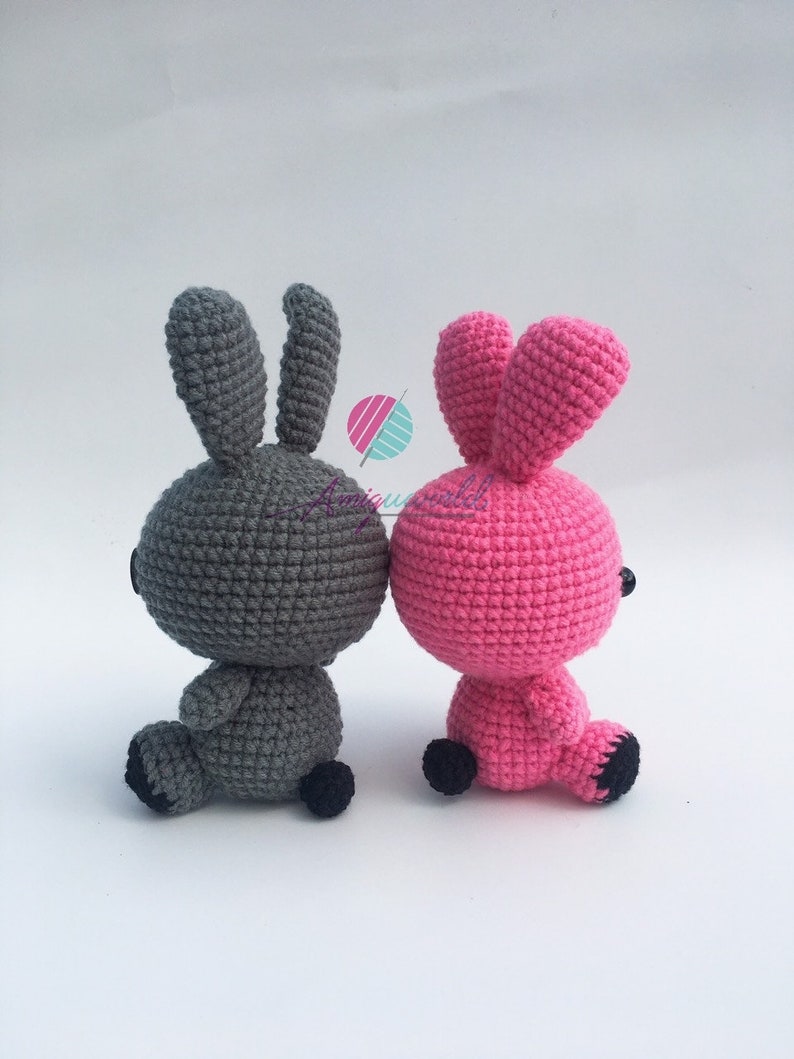 May include: Two crocheted bunny plushes, one grey and one pink, sitting back to back. They have black eyes and black feet.