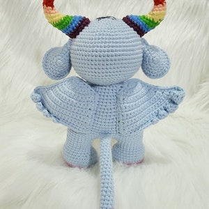 May include: A light blue crocheted stuffed toy with rainbow horns, wings, and a pink tail. The toy is standing on a white fluffy surface.