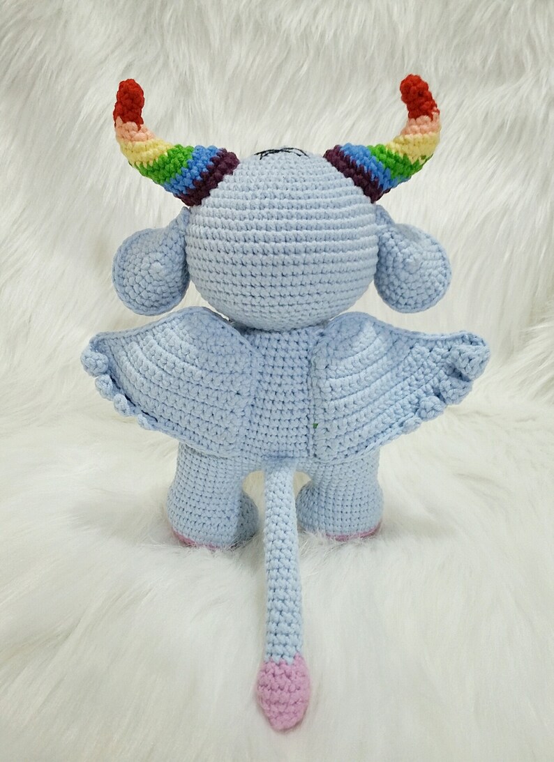 May include: A light blue crocheted stuffed toy with rainbow horns, wings, and a pink tail. The toy is standing on a white fluffy surface.