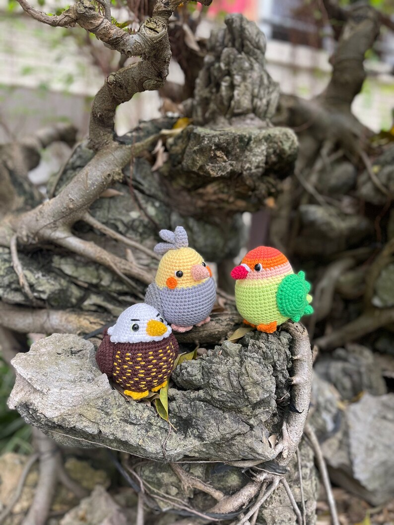May include: Three crocheted bird figurines, a brown and white eagle, a yellow and gray cockatiel, and a green and orange lovebird, are perched on a tree branch.