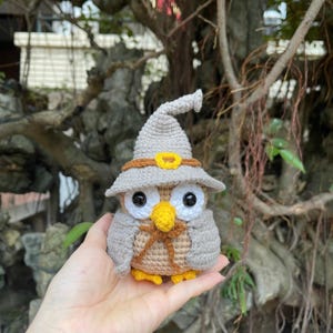 Puede incluir: Un búho de ganchillo hecho a mano, con un sombrero de bruja gris con borde marrón y hebilla amarilla. El búho tiene grandes ojos negros, un pico amarillo y está en tonos de gris, marrón y amarillo. Un artículo artesanal caprichoso.