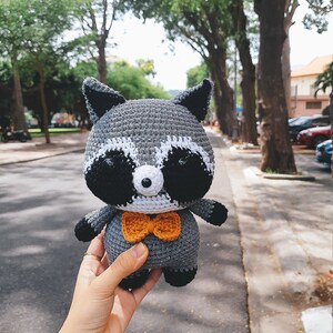 May include: A crocheted raccoon plush toy wearing a bow tie. The toy is grey with black eyes and a white nose. The bow tie is orange.